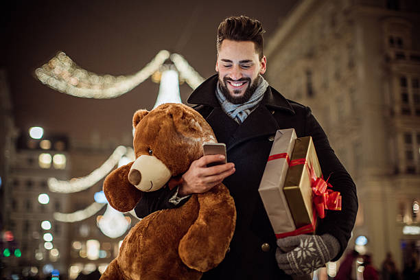 Can't wait to see her face Young man holding Christmas gifts and big Teddy bear. Checking his smart phone. Wearing warm clothing. Vienna, Austria. man with teddy bear stock pictures, royalty-free photos & images