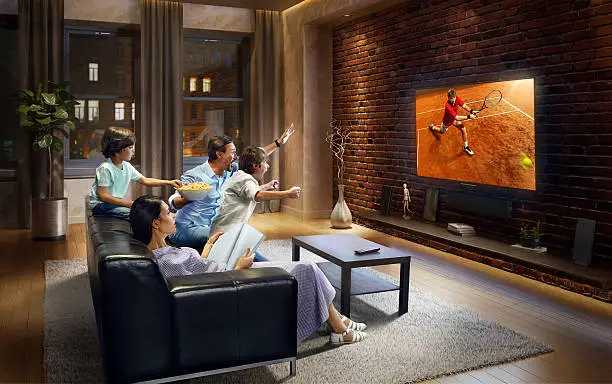 :biggrin:Father and two young children cheering and watching Tennis game on TV. Mother is reading a magazine. They are sitting on a sofa in the modern living room. The TV set is on the loft brick wall. It is evening outside the window.