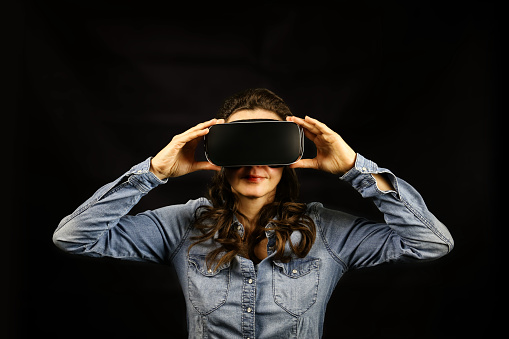 Single young woman holding large 3D virtual reality viewing device over face on black background