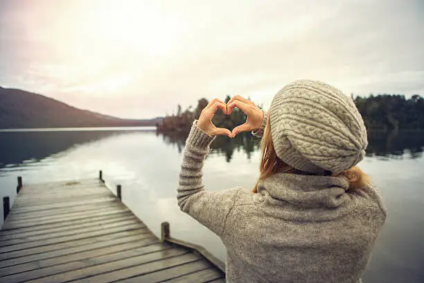 Photo of Young woman on lake pier, makes heart shape finger frame