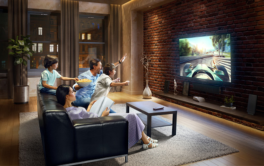 :biggrin:Father and two young children cheering and watching Sports Car Race on TV. Mother is reading a magazine. They are sitting on a sofa in the modern living room. The TV set is on the loft brick wall. It is evening outside the window.