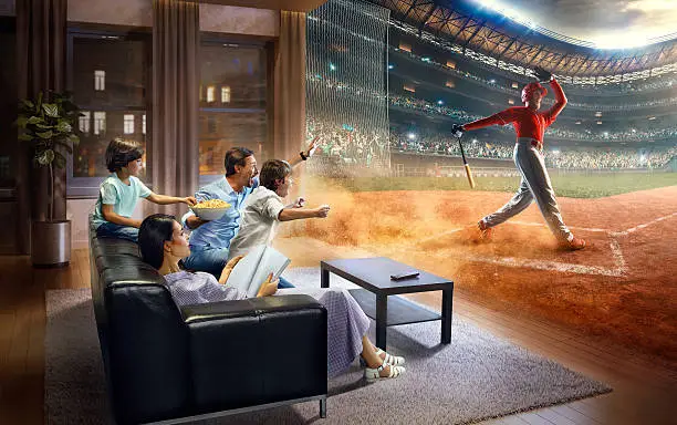 :biggrin:Father and two young children cheering and watching extremely real Baseball game. Mother is reading a magazine. They are sitting on a sofa in the modern living room faced to a real stadium. It is evening outside the window.