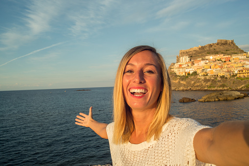 Young woman in Italy takes a selfie portrait with Castelsardo village and seascape on the background. Sunset light, late summer day.