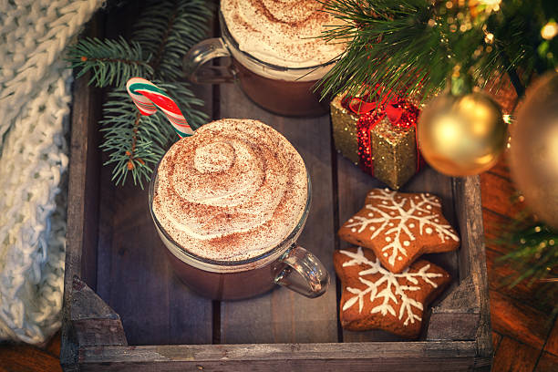Hot chocolate with whipped cream and candy cane in old wooden basket near Christmas tree. There are also gingerbread cookies and Christmas decorations