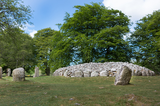 Inverness, Scotland - June 2, 2012: The menhir stone circle and grave site heap of gray stones at prehistoric Clava Cairns. Surrounded by green trees, grassy field and white clouds in blue sky.