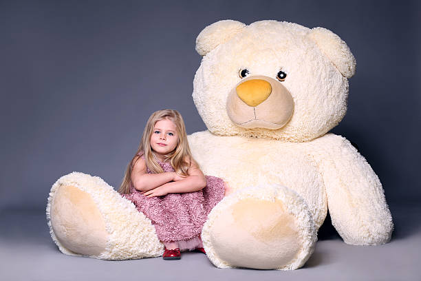 Little model in dress sitting on the big teddy's Little model in dress sitting on the big teddy's leg, stuffed animals, plush animals, big plush toys, kids and gifts, lilac dress, blonde girl, baby in studio, cerne abbas giant stock pictures, royalty-free photos & images