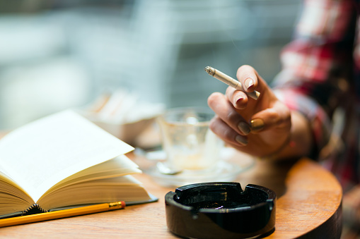 Open book and yellow pencil, ashtray and female hand with cigarette. Shallow DOF, focus on hand