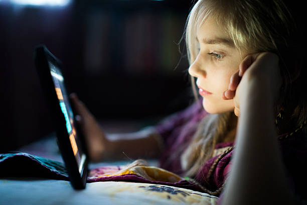 Teenager girl reading from tablet at night in the bed Beautiful long haired teenager girl reading from tablet, in the dark room at night in the bed, with her face illuminated by the screen 13 14 years child teenage girls lying down stock pictures, royalty-free photos & images