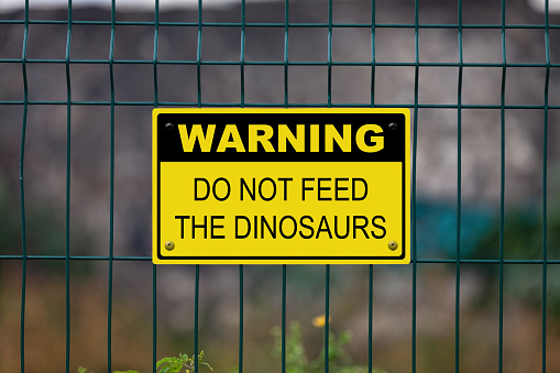 Black and Yellow warning sign attached on a fence. The sign stating “Warning - Do not feed the dinosaurs”.