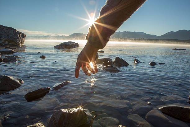 Finger touches surface of mountain lake. The sun and the landscape are reflecting on the water.