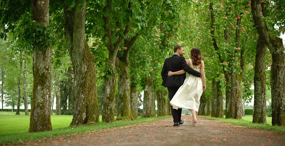 Bride walking with groom on single track in park.