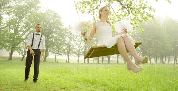 Groom pushing bride on swing in park.