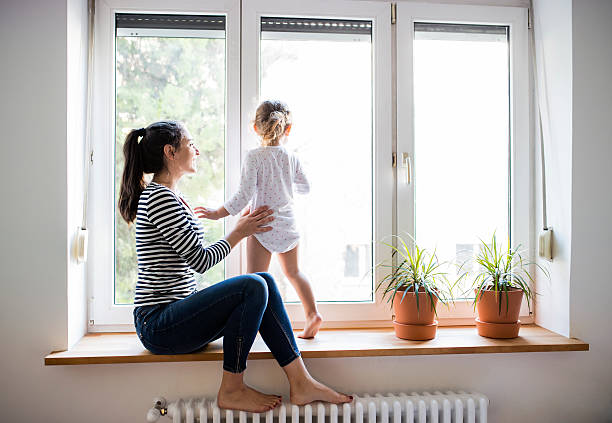 Mother with her little daughter looking out of window Beautiful young mother sitting on window sill with her cute little daughter looking out of window jeans photos stock pictures, royalty-free photos & images
