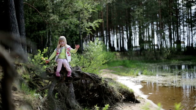 Pretty blond girl sitting on an old tree stump with roots on the bank of the lake.