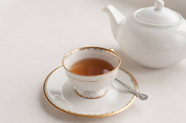 An old fashioned (vintage) cup of tea and a silver spoon with a white tea pot in the background. It is a brew of white tea but still it has a nice dark tone and matches the golden lining of the cup and the saucer, you can see the handle of a silver spoon peeking out behind the cup.