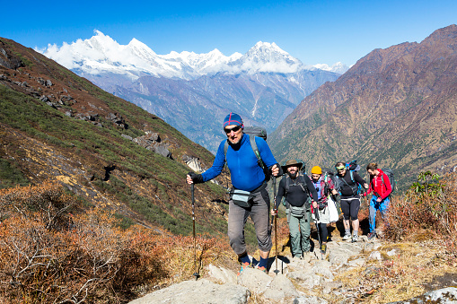 Group of People of different age and ethnicity walking up on Mountain Trail during Hike in Nepalese Himalaya carrying heavy backpacks and climbing gear led by mature Guide