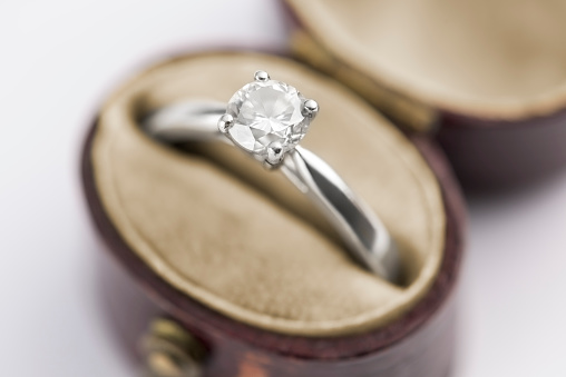 A sparkling solitaire diamond ring is in vintage red leather ring box on a velvet cushion. The 1-carat diamond is set on a platinum band. The ring box is on a white background.