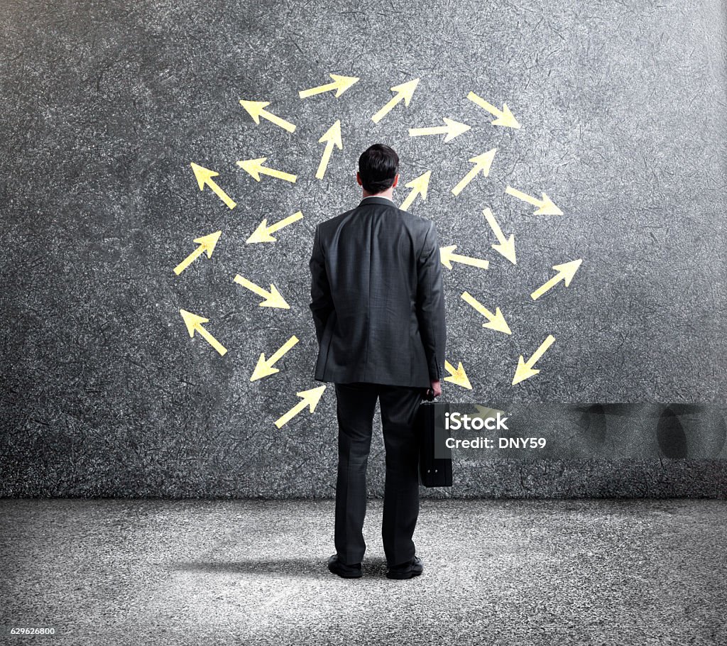 Businessman Looking At Arrows Pointing In Different Directions A rear view of a businessman standing while holding his briefcase as he looks up towards a wall with arrows pointing in different directions. 30-34 Years Stock Photo