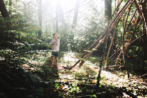 A depiction of healthy childhood play on a warm summer day.  A cute boy has fun as he plays in and builds a teepee fort in the forest out of fallen branches, using his imagination for adventure and outdoor discovery.  Horizontal image with copy space.