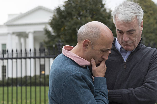Gay Couple Looking Distraught In Front of White House Mature, older, senior gay male couple looking unhappy and distraught in front of White House. sad gay stock pictures, royalty-free photos & images