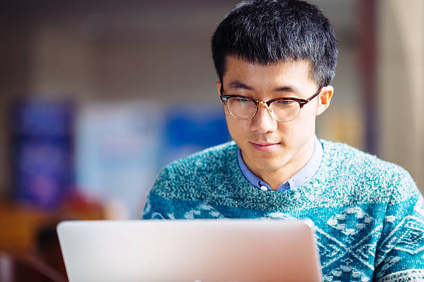 young asian man student using laptop young asian man student with sweater using laptop in library male chinese student stock pictures, royalty-free photos & images