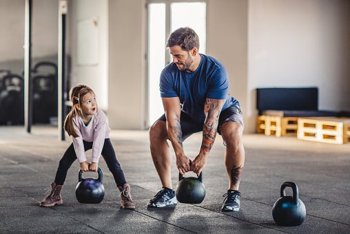 Father doing his training with kettlebells in gym while his little daughter supporting him.