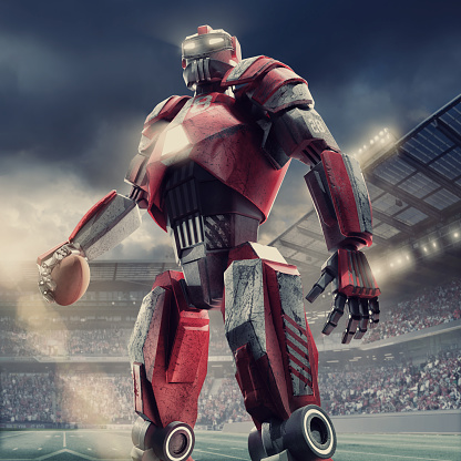 A low angle view of a red and white American football robot covered in scratches, scuffs and dents, standing with arms apart holding an American football. The cyborg is standing in an American football field in a generic outdoor floodlit stadium, full of spectators under stormy evening sky.