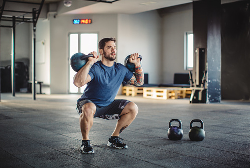 Youg man doing squats with kettlebells at gym. Professional athlete. Wearing sports clothing.