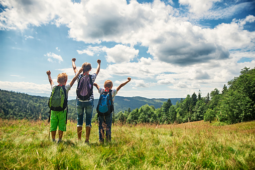 Three happy kids hikers after reaching the hill top. The girl and her two brothers are rising arms in gesture of success.