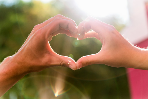 A photo of couple forming heart with human hands. Cropped image of man and woman making shape against shiny sun. Image is depicting love.