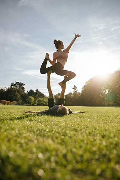 Healthy young couple doing acrobatic yoga Vertical shot of healthy young couple doing acrobatic yoga on grass. Man and woman exercising in pair at the park. acroyoga stock pictures, royalty-free photos & images