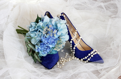 Bright shoes for stylish bride. Bright blue suede high hill pair of shoes and brides bouquet with blue flowers on a white bridal veil. Wedding background.