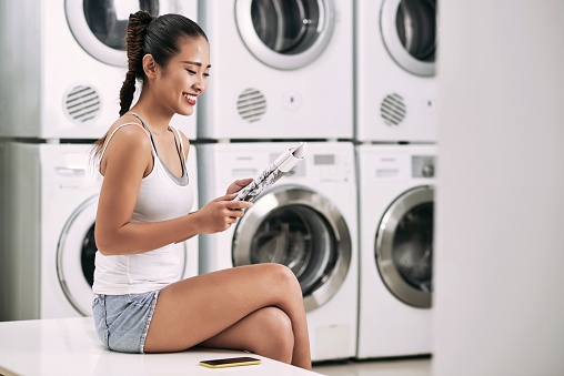 Smiling young woman enjoying reading magazine in laundry room