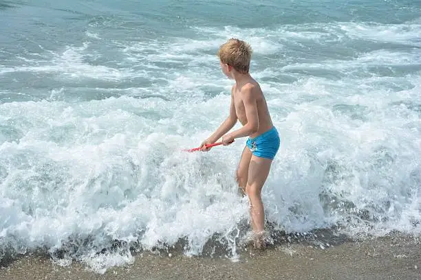 Small blond boy with blue swimming trunks stands with a fishing-net directly in the sea - wave
