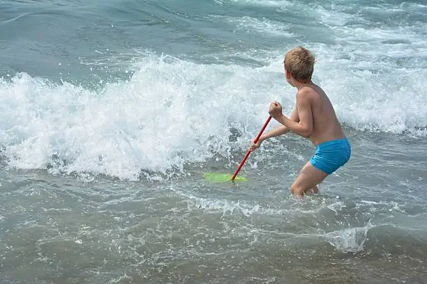 Small blond boy with blue swimming trunks stands with a fishing-net in the water and waits for the sea - wave