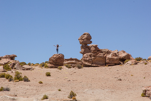Potosi, Bolivia - October 30, 2016: Man with curly hair, wearing a grey long-slleve shirt and blue pants,  doing the karate crane position, on the top of rock, in a desertic landscape.