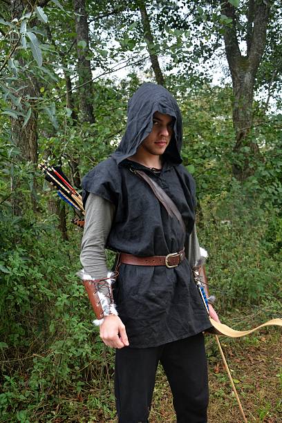 Medieval archer with black hood and coloured arrows in the quiver stands with bow  in the nature and looks forwards