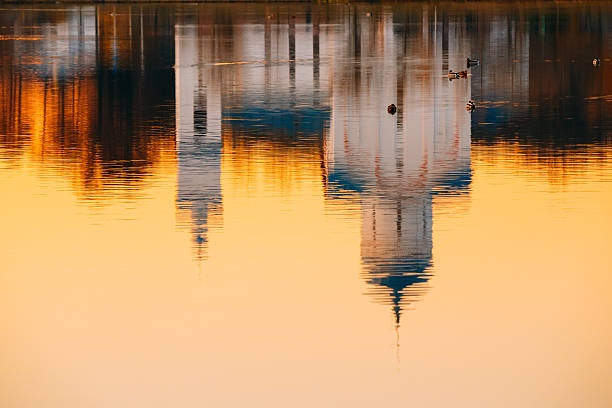 The Reflection Image Of Russian Orthodox Christian Temple Church Chapel On Sunset Sunrise Dawn River Lake Water Surface, Russia. A Small Flock Of Floating Ducks Back From.