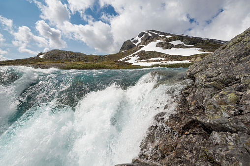A wide angle (14mm lens on full frame DSLR) view of the mountainous landscape of Jotunheimen National Park in Norway. A stream of water (or a small river) - from melting snow and ice - rushes down stairs of rocky ground and boulder making a waterfall with foam and splashing water on a warm sunny day in the summer (July). The winter season is long here and all snow and ice may not melt until July or August - or not at all. The mountainsides are covered with fresh green grass, ferns and bushes. Some patches of snow are still present. The sky is blue with white puffy clouds.