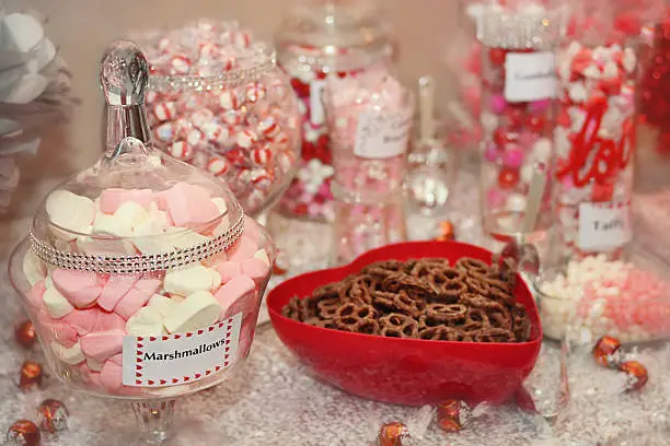 A table with various types of candies, marshmallows and chocolate covered pretzels. Sweets are red, pink and cream colored.