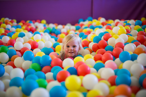 A little blond girls plays in a ball pool full of multi colored balls. She is excited and happy