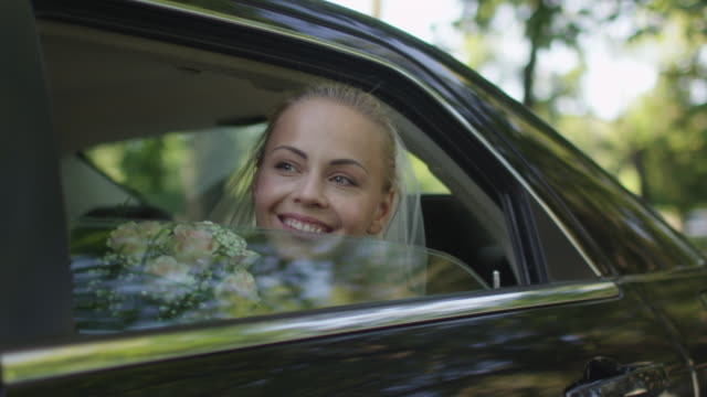 Young beautiful happy bride sits in a car and waves out of the window.