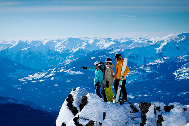 Group of skiers on top of mountain pointing at valley. Scenic photo of group of friends standing on edge with skis and pointing across valley. Snowy mountain range in background top of ski mountain stock pictures, royalty-free photos & images
