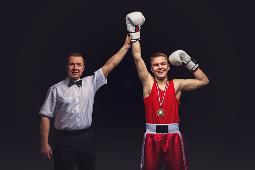 Boxing referee gives medal to young teen boxer in red form and white gloves. Winner. Studio shot on black background. Copy space.