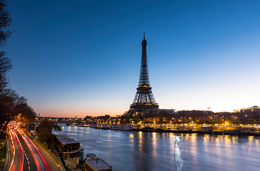 An early morning sunrise takes place behind the majestic Eiffel Tower in the city of Paris in France. Situated near the Seine river, the Eiffel Tower is one of the most important monument in the World. On the left side, cars are on the move and leave some trail lights.