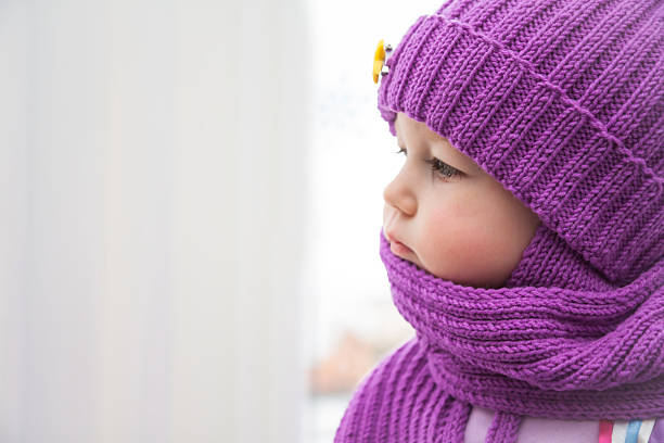 Sad child looking into the window during cold winter day in warm clothes