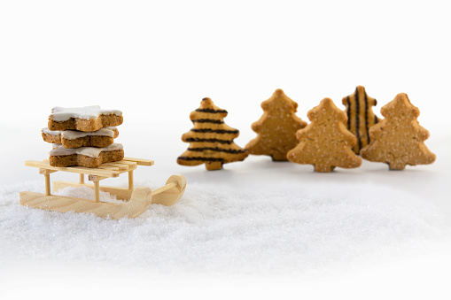 Wooden sled carries cinnamon star cookies through the snow in front of christmas trees made from gingerbread, background with copy space fades to white, selective focus, narrow depth of field