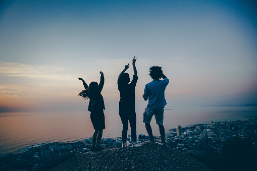 Group of multi-ethnic friends partying and dancing outside on a rock at sunset on summertime watching the city