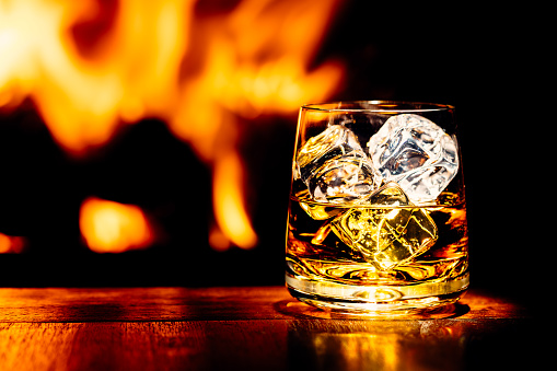 Glass of whisky with ice on a wooden table, with flames from a wood burning stove in a dark background.