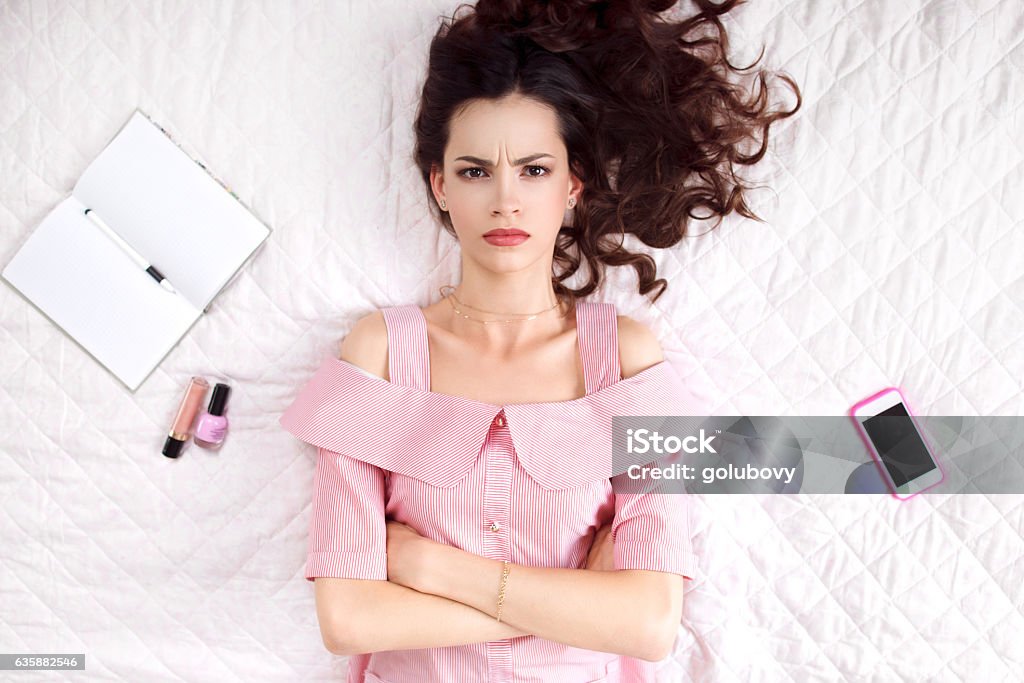 Pouted woman lying with crossed hands top view Pouted woman lying with crossed hands on bed top view. Resentful brunette in pink looking at camera, ready to cry. Offence, caprice, feeling concept Women Stock Photo